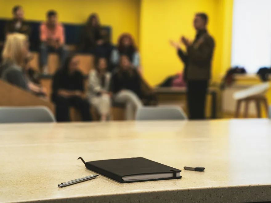 Un carnet posé sur une table, devant un public qui écoute un intervenant